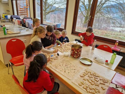 Kinder sitzen an einem Tisch und verzieren Plätzchen in einem Raum mit großen Fenstern und Blick auf Bäume