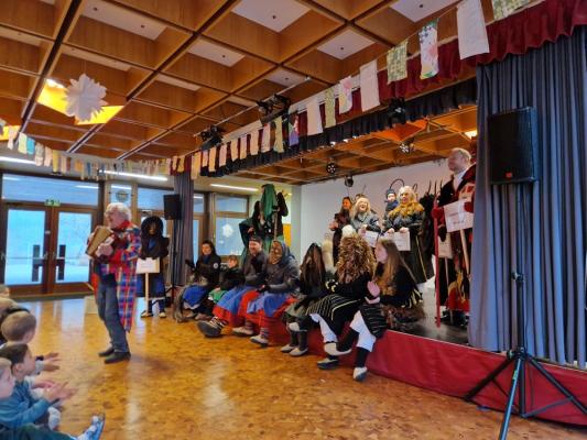 Mehrere verkleidete Personen mit Umhängen, Hüten und Masken sitzen und stehen auf einer Bühne in einem geschmückten Saal, während eine Person mit kariertem Mantel und Akkordeon davor steht.
