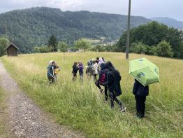 Eine Gruppe von Personen steht in einem hohen, grünen Grasfeld vor einem bewaldeten Berghang.