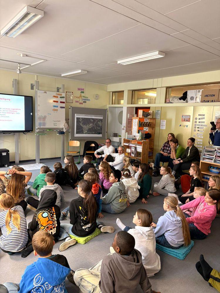 Gruppe von Kindern sitzt auf dem Boden eines Klassenzimmers und schaut auf eine Präsentation an der Wand
